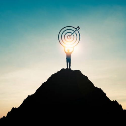 Silhouette of businessman holding target board on the top of mountain with over blue sky and sunlight. It is symbol of leadership successful achievement with goal and objective target.